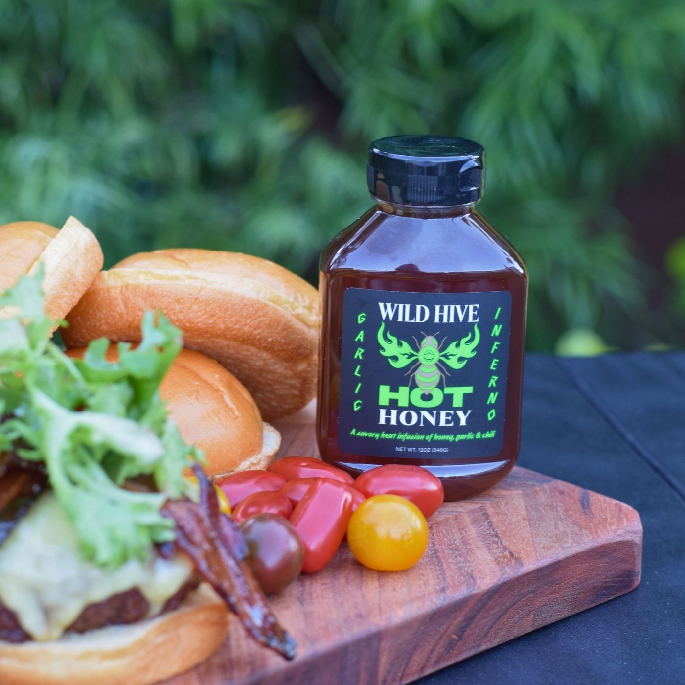 Bottle of Wild Hive Hot Honey Garlic Inferno on a cutting board with a burger and vegetables outdoors.