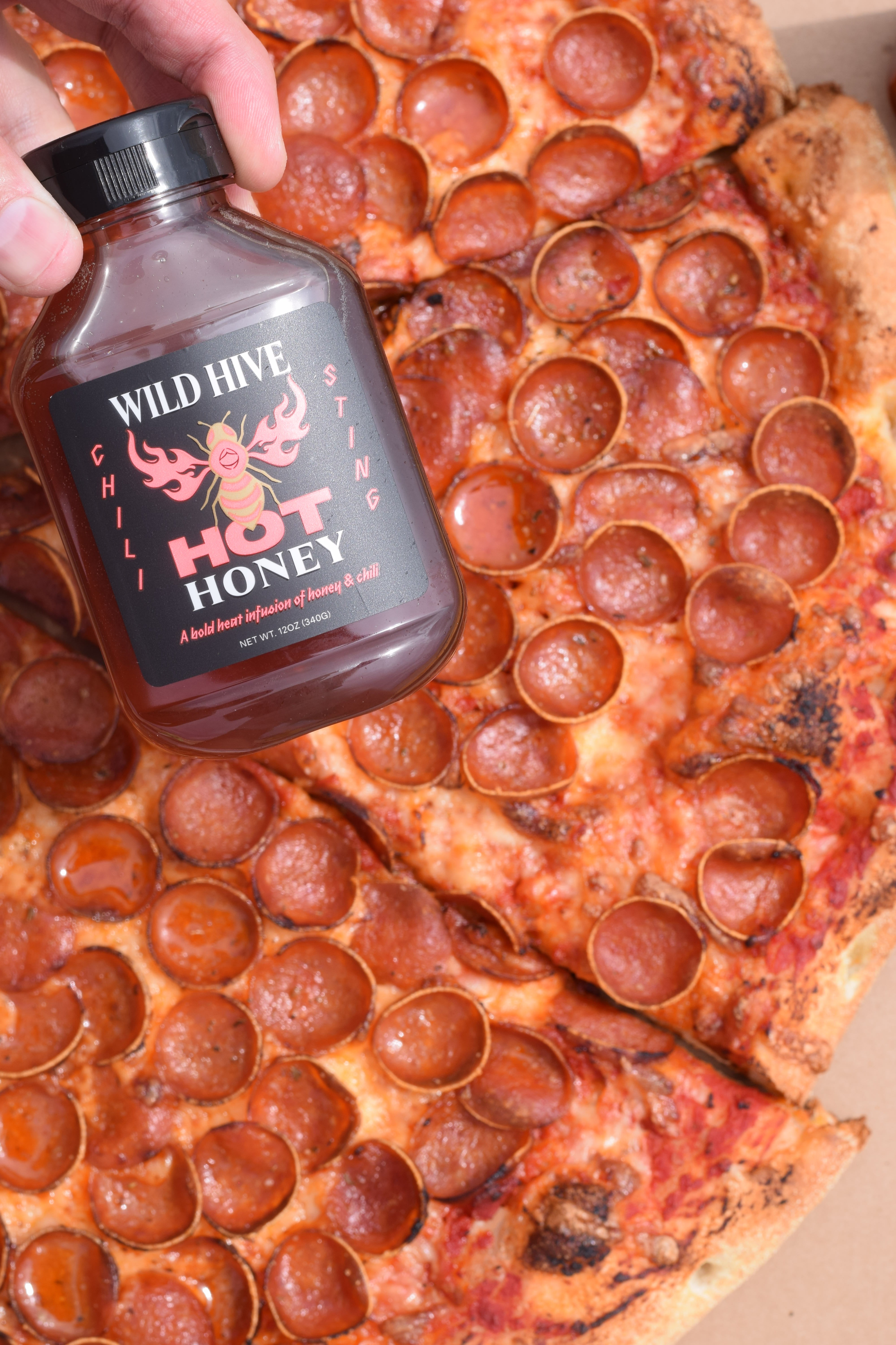 Hand holding a bottle of Wild Hive Hot Honey over a pizza with pepperoni slices.